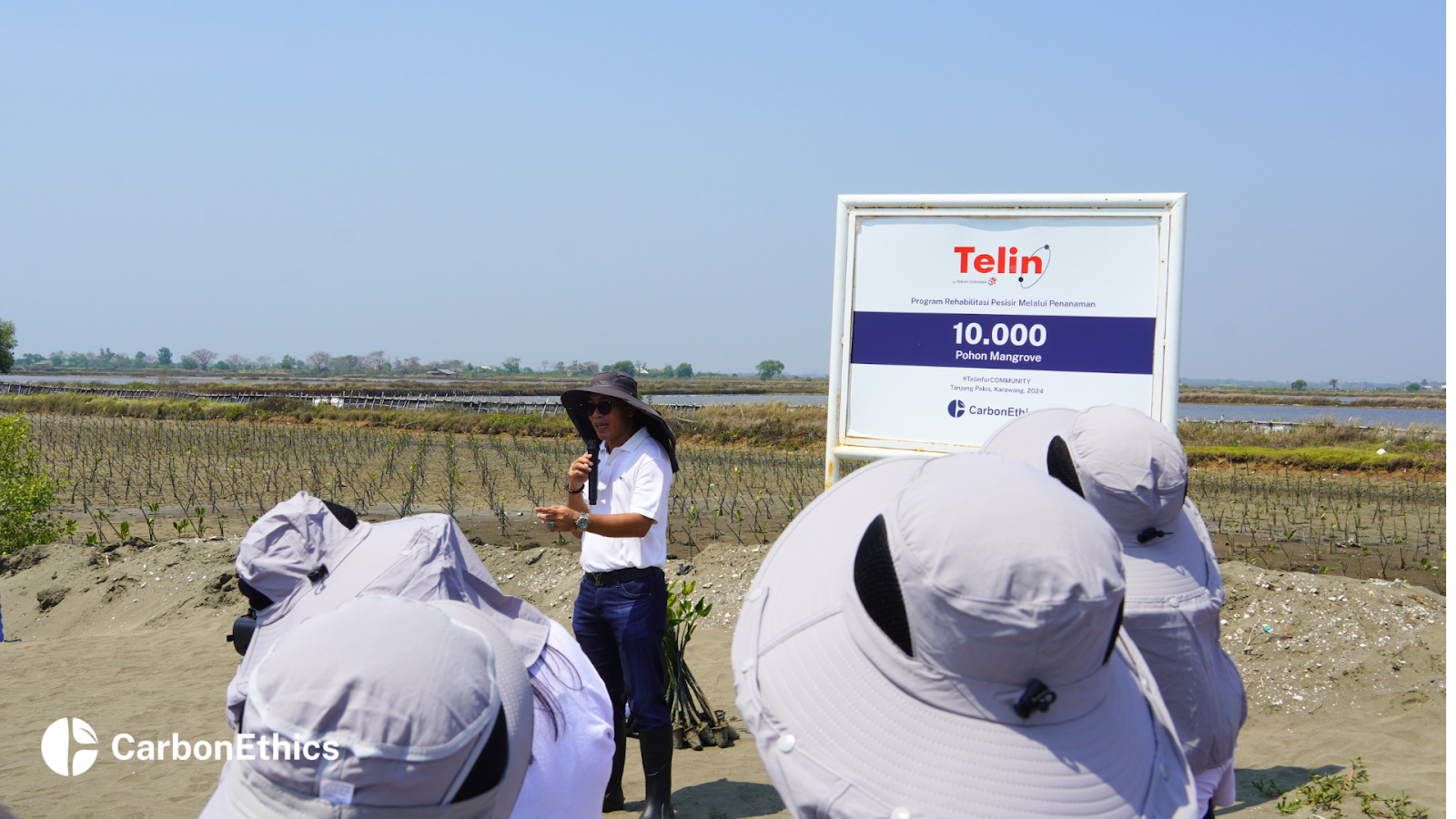 A representative giving a speech during the 10,000 Mangrove Planting Expansion event, with attendees wearing hats in the foreground and the Telin project signboard in the background at the mangrove restoration site in Tanjungpakis, Karawang, West Java.
