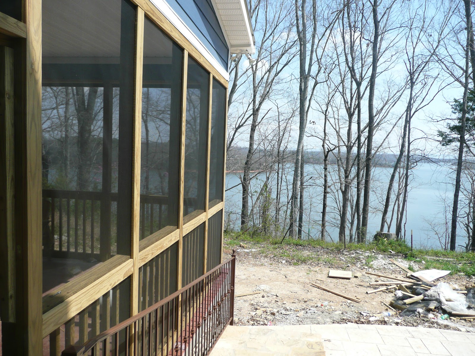 View from a screened-in porch with wood framing and a metal railing, overlooking a lake and a wooded shoreline during construction.