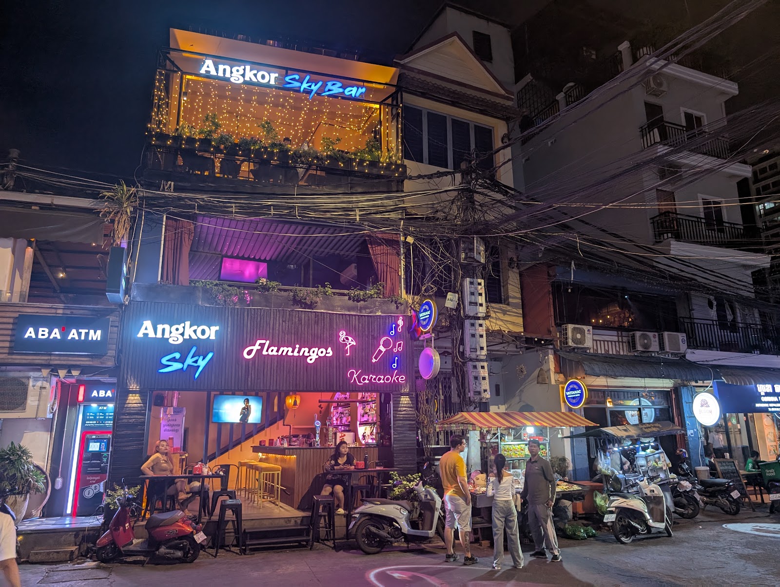 Cables converging on a telephone pole in front of an apartment
                 building amid lit up neon signs for businesses on the street
                 level