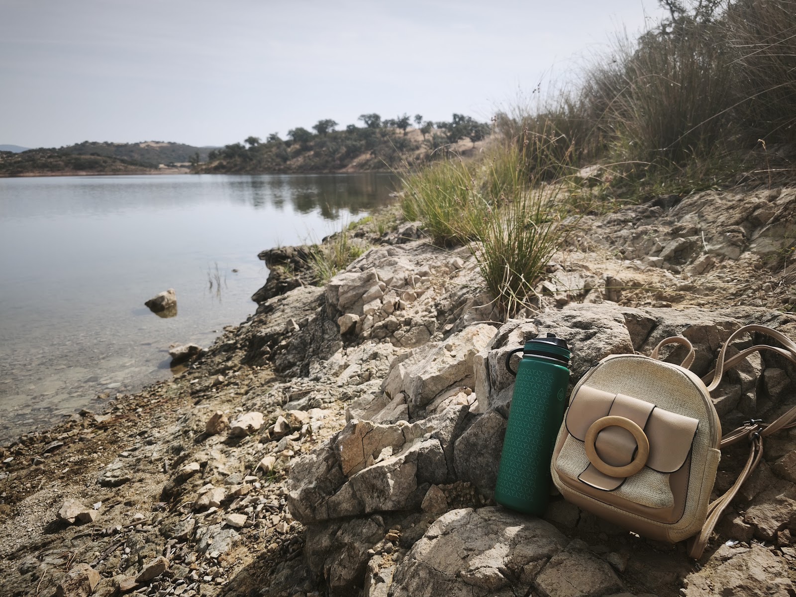A backpack and reusable water bottle resting on rocks beside the Embalse de Zufre reservoir before a summer swim.