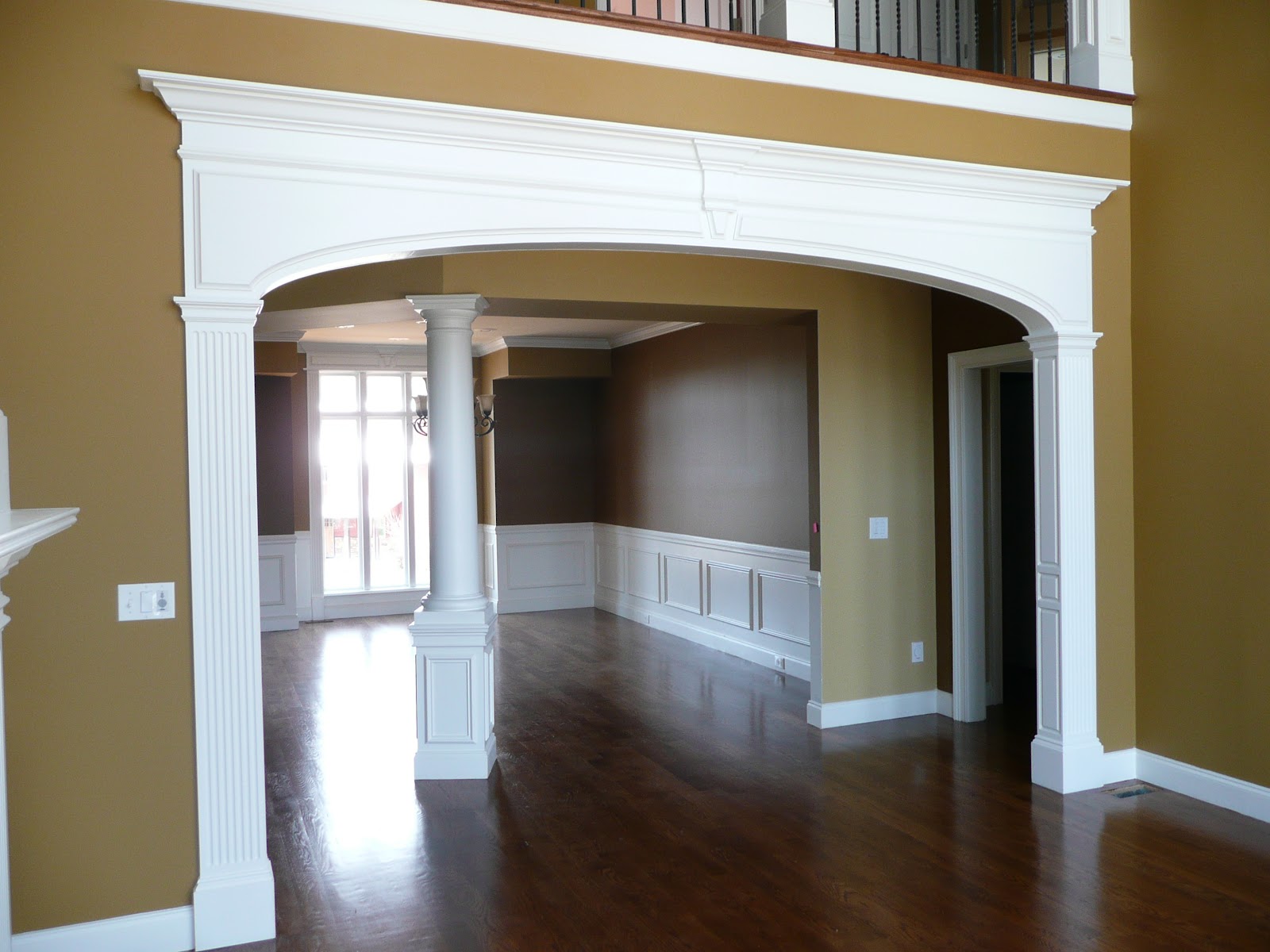 Interior view of a house featuring a white arched doorway with fluted columns, dark wood flooring, tan walls, and white wainscoting.