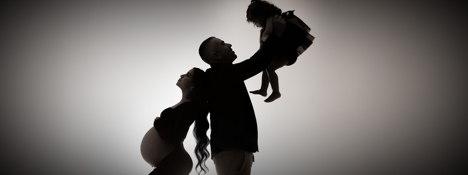 Silhouette maternity portrait: pregnant mother back-to-back with partner lifting their little girl in a studio