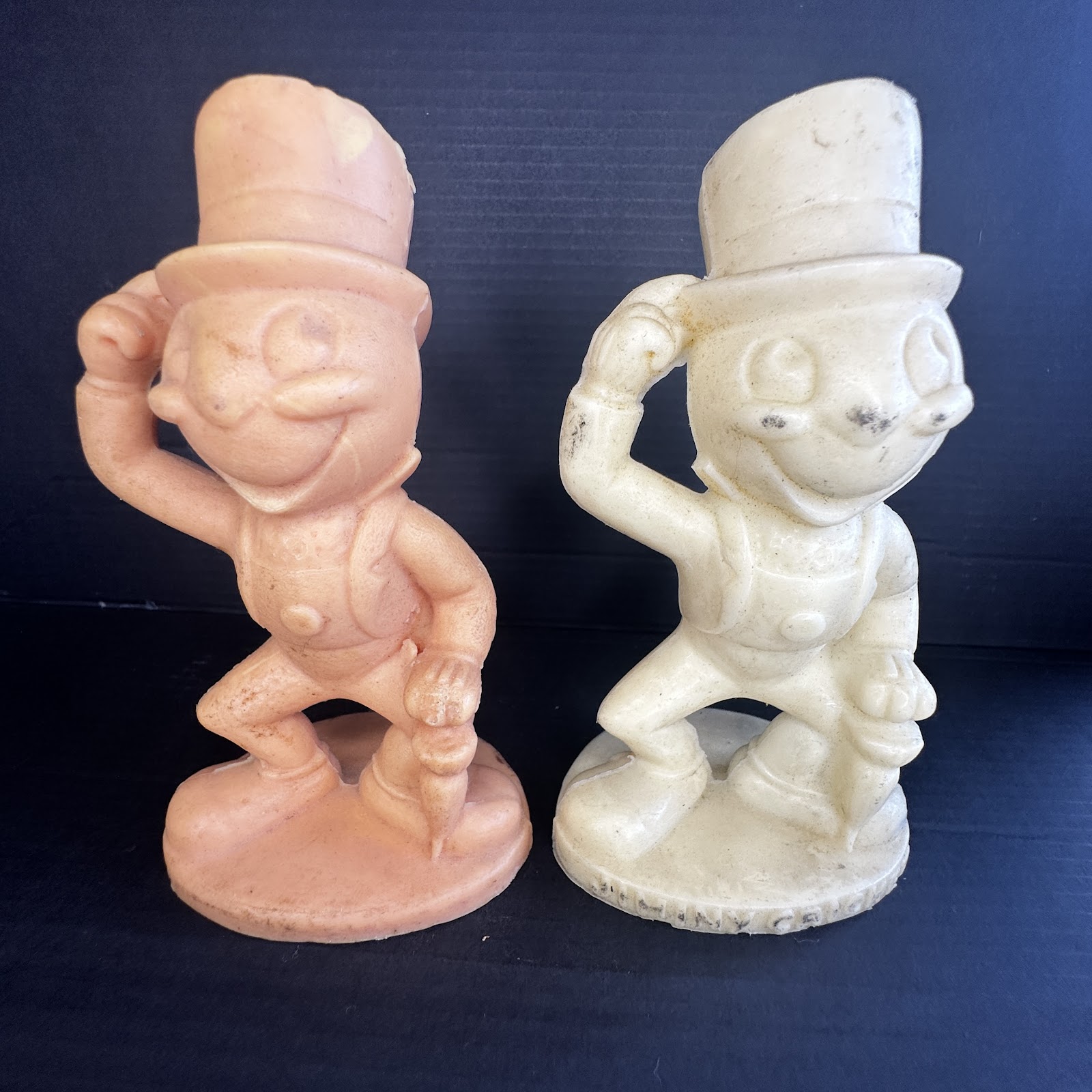 Pair of 1950s Sun Rubber Co. Jiminy Cricket vulcanized rubber squeak toys, one peach-toned and one cream, each in hat-tipping pose with umbrella on circular embossed base