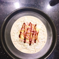Trải bánh tortilla trải lên mâm nướng, tiếp theo cho bắp cải và trải đều mặt (nhớ là không nên cho ra ngoài bìa bánh). Thêm mozzarella vào giữa bánh + xúc xích cắt sợi + trứng gà đánh tan và cuối cùng là xịt sốt teriyaki lên trên bề mặt để bánh được thấm và ngon hơn.