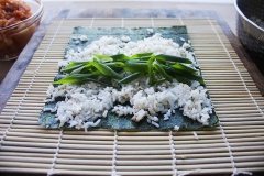 Dùng mành tre cuốn kimbap mua ngoài chợ đặt lên mặt phẳng, trải 1 tấm rong biển lên trên, múc cơm vào dàn đều khoảng 4/5 tấm rong biển, xếp 1 lớp hành lá vào 2/3 cơm. Kim chi vắt ráo nước, cắt nhỏ, xếp trên hành.