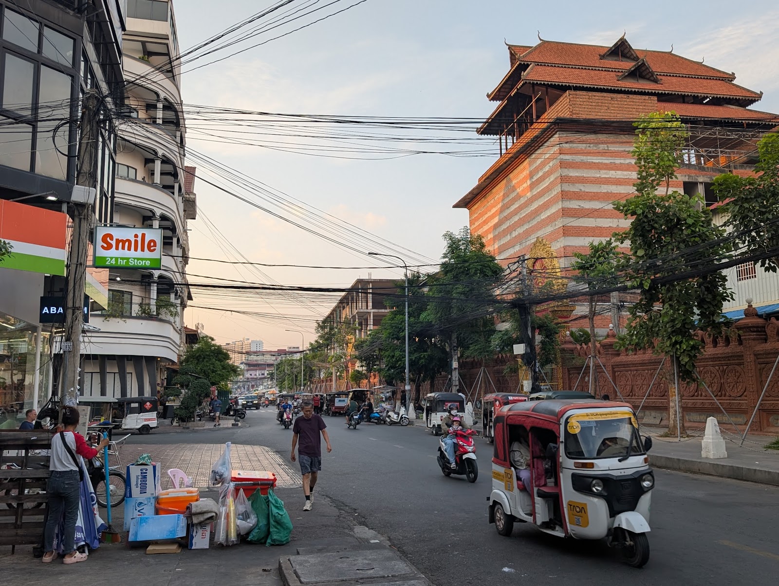 A busy street corner in Phnom Penh with some cables
                 zigzagging in front of a sky near sunset above
