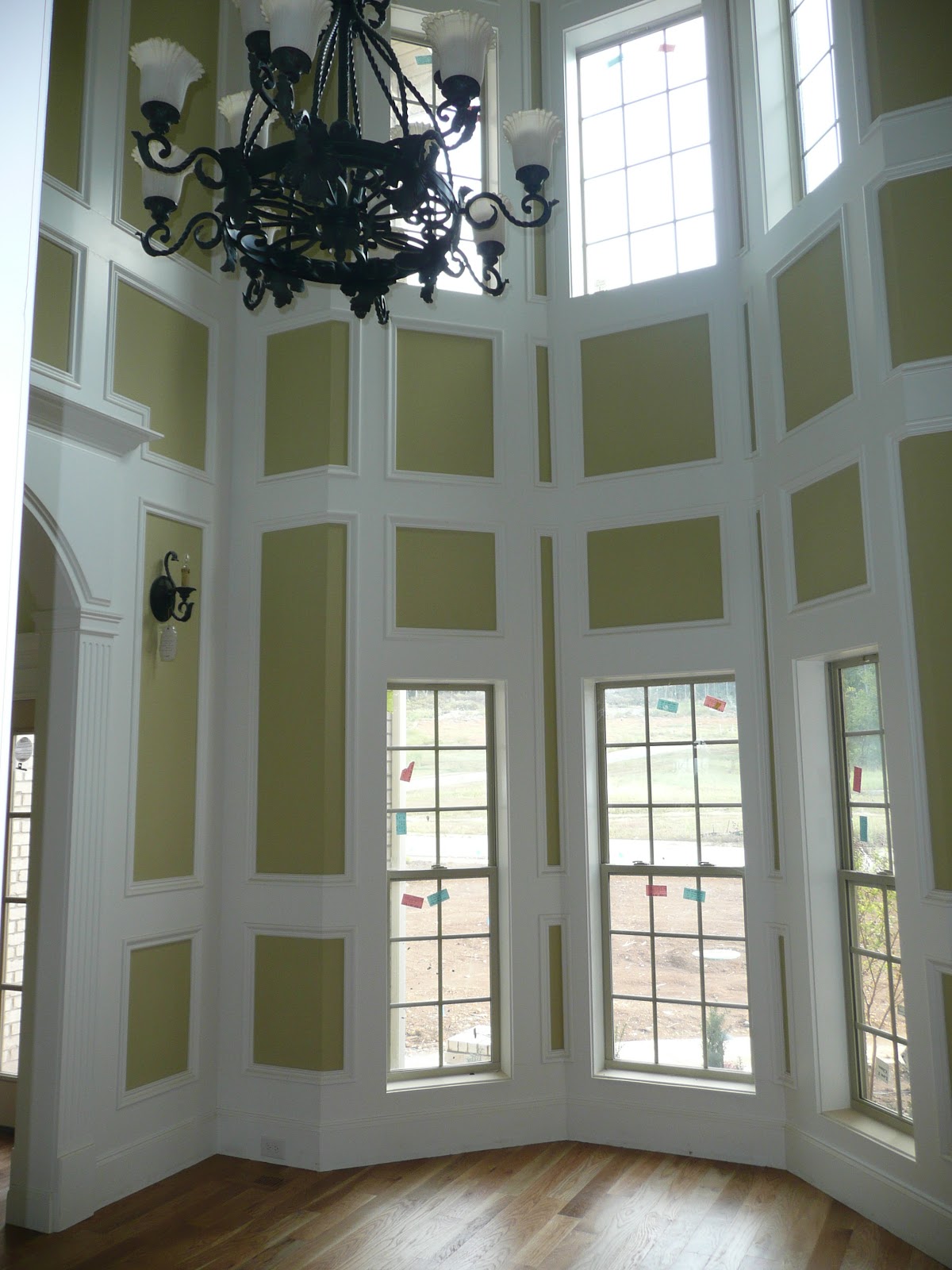 High-ceilinged interior room with tall windows, white decorative wall paneling on tan walls, and an ornate black chandelier.