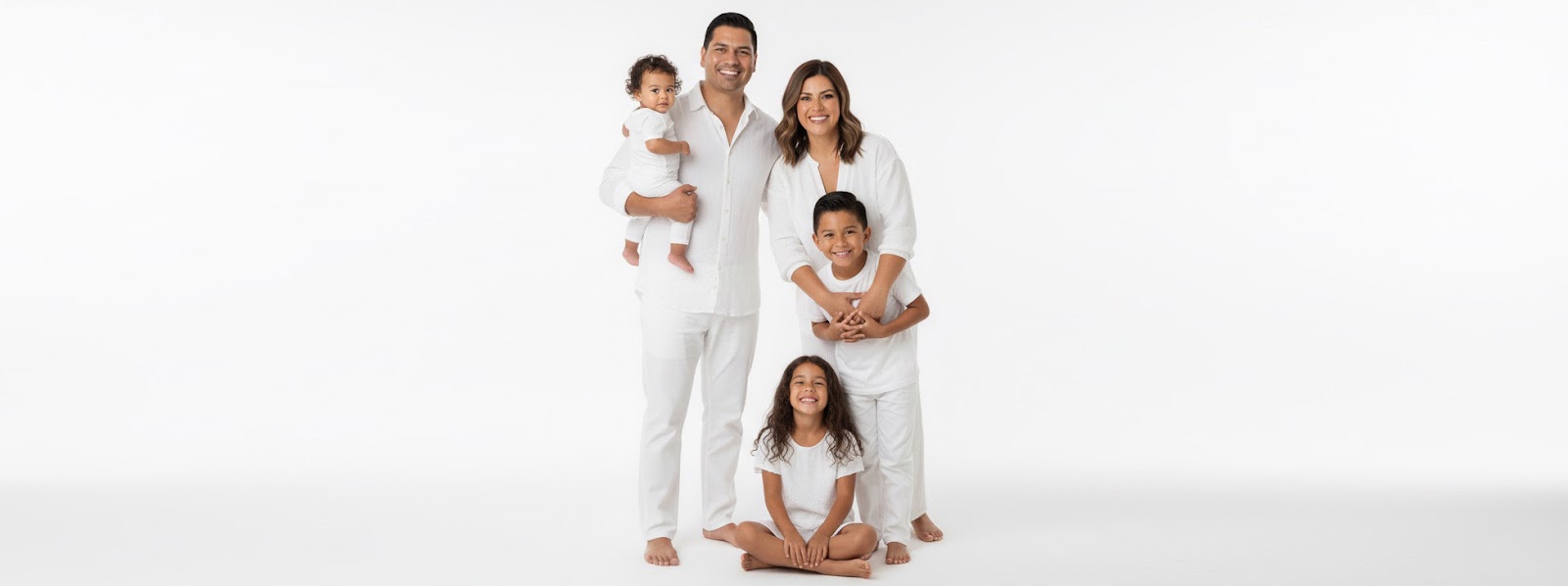 Family of five in white outfits on a white studio background—parents standing with toddler, older son in front, daughter seated smiling.