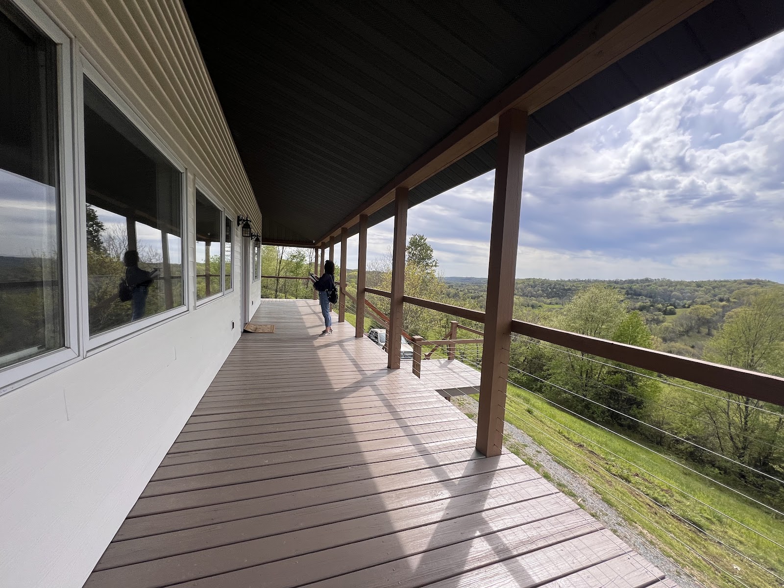 A long wooden deck with a railing overlooks a scenic valley of green trees. The house features white siding and large windows reflecting the outdoor view.