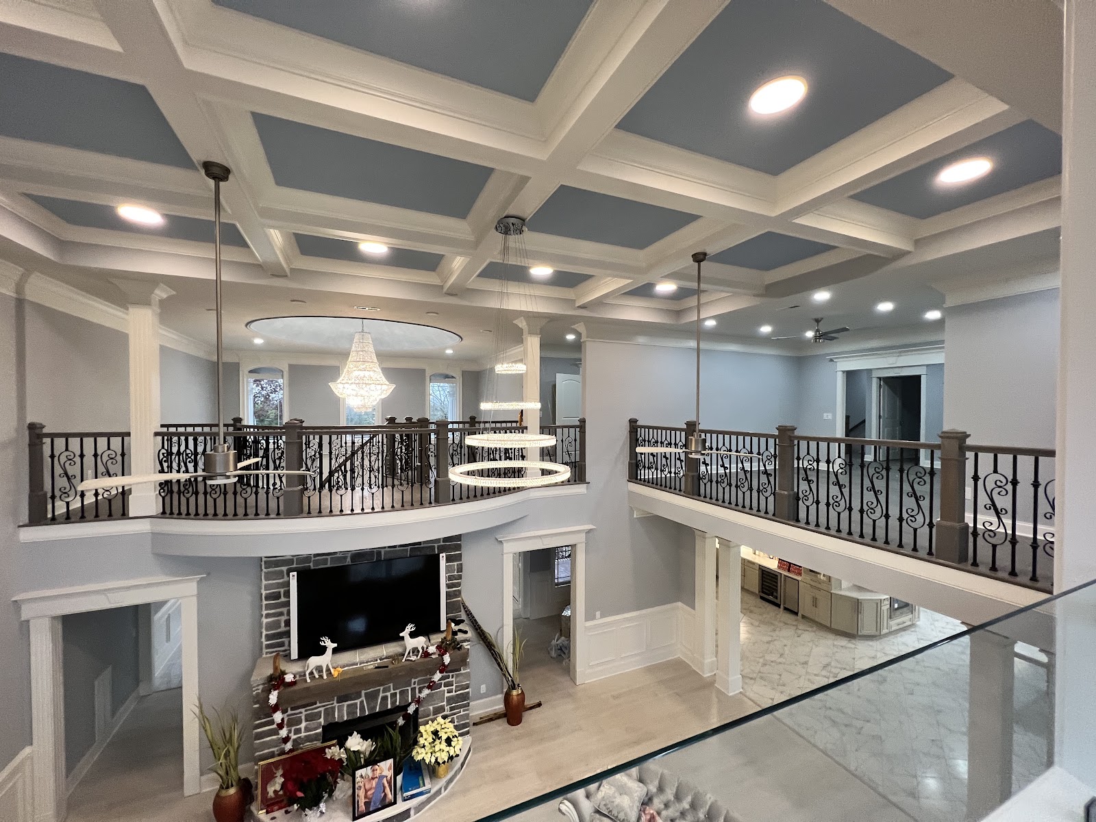 High-angle view of a residential interior featuring a coffered ceiling with blue panels, a curved second-story balcony with wrought iron railings, and a stone fireplace.