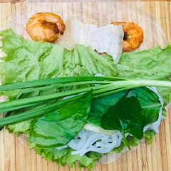 Trải bánh tráng, dùng rau xếp vào, cho thêm bún và các loại rau sống, thêm 2 con tôm và 2 miếng thịt ở góc cuối. Dùng tay cuốn chặt, tiếp theo cho hẹ vào là xong. Lần lượt làm cho hết phần thịt và tôm.