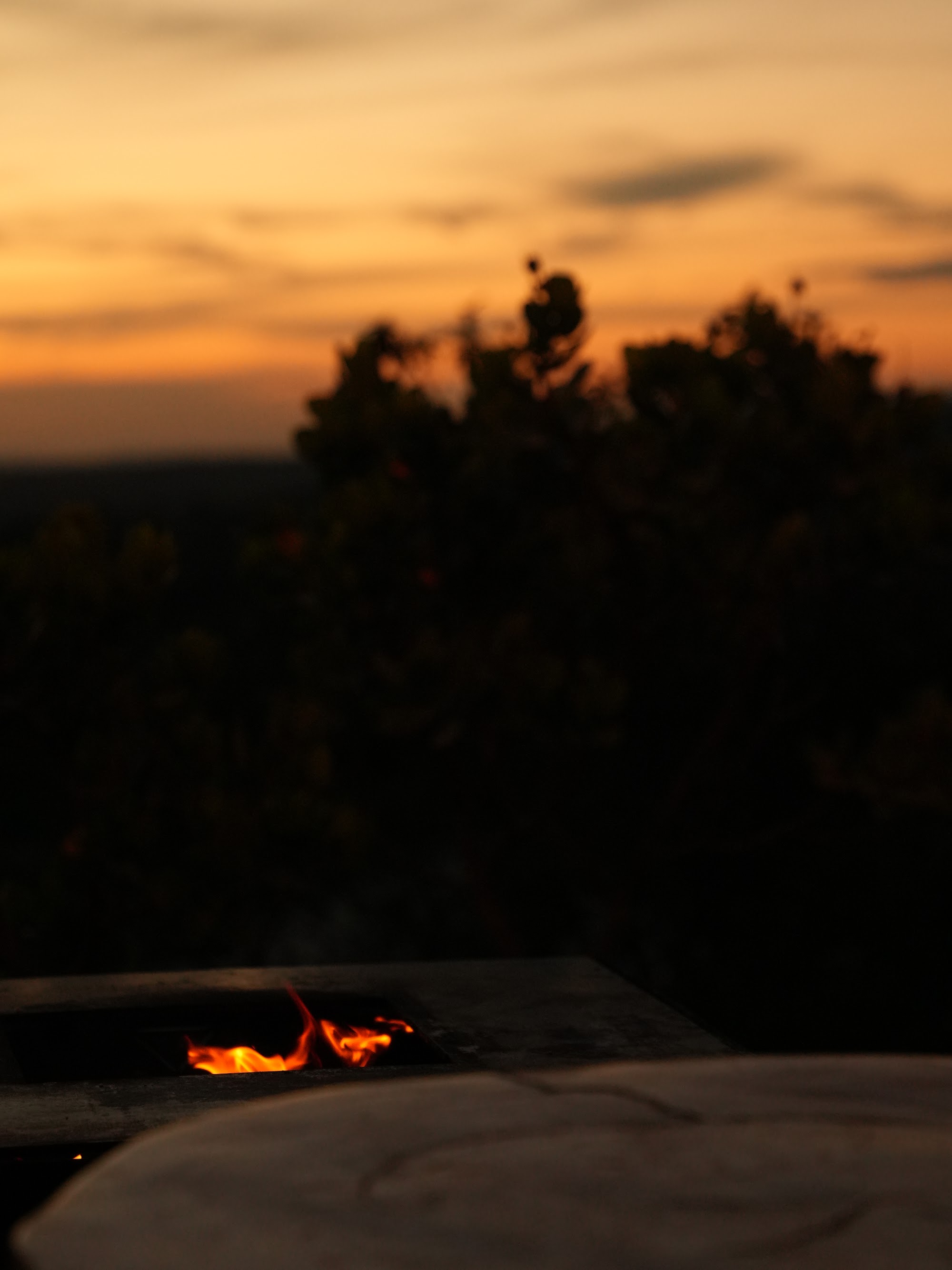 Detalhe do pit fire aceso no deck de um chalé Mei do Mato ao entardecer, com o céu em tons de laranja ao fundo