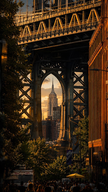 Looking through a massive steel bridge tower arch to reveal a distant art deco skyscraper framed perfectly at golden hour.