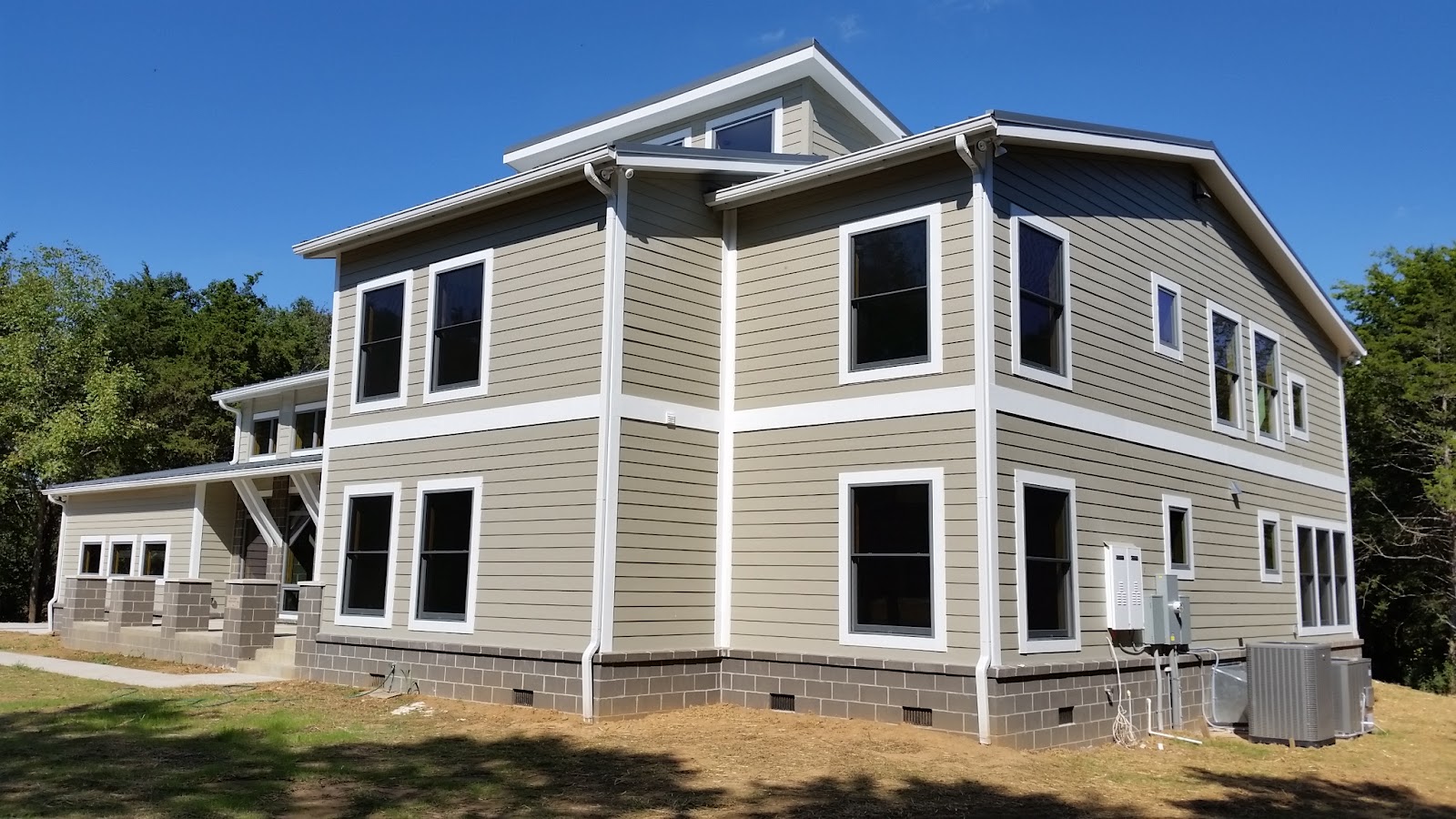 Side exterior view of a residential building showing grey siding, white trim, multiple windows, and exterior HVAC equipment.