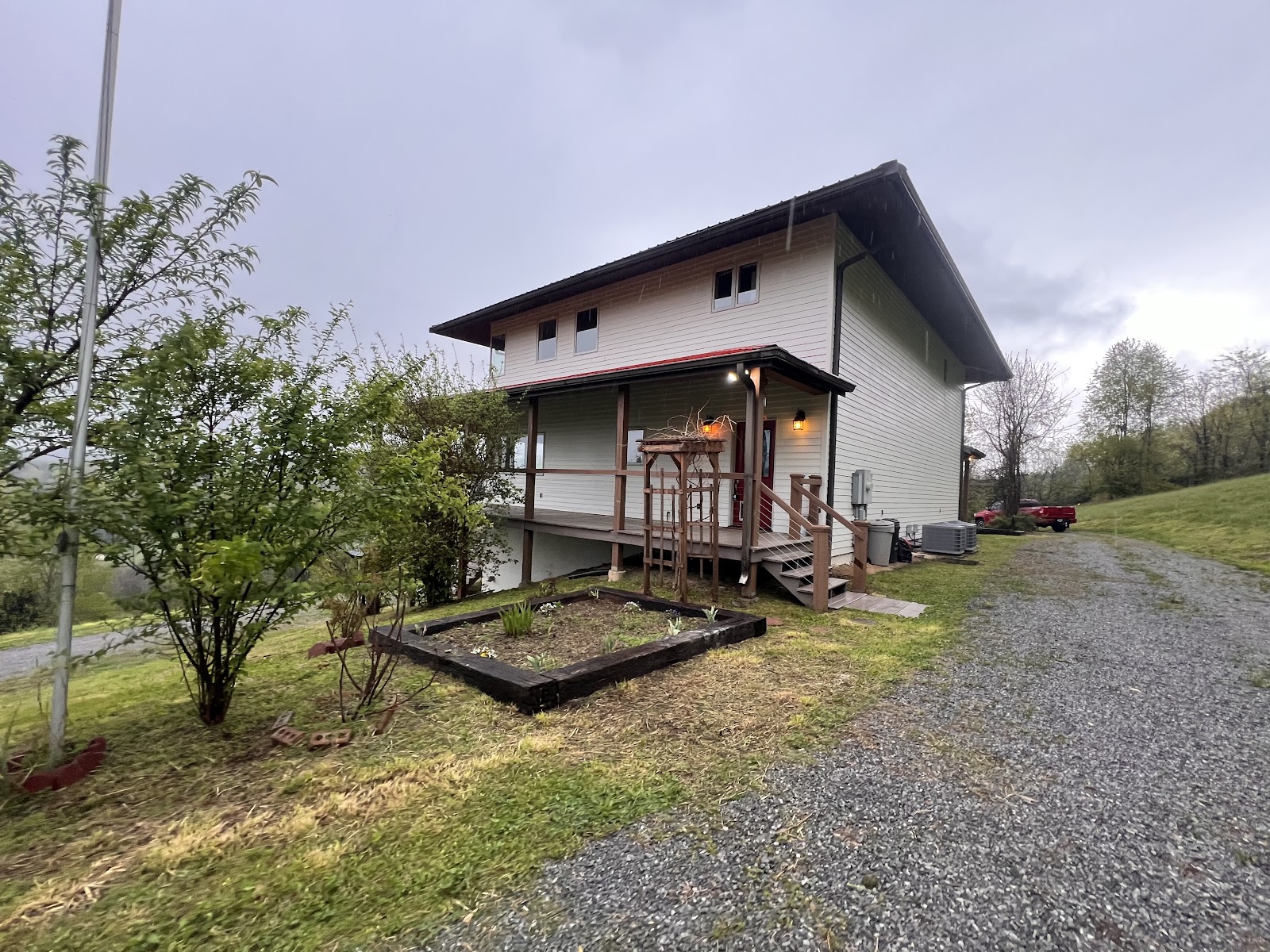 A two-story house with light siding and a dark roof, featuring a wooden porch and deck. A gravel driveway and a small garden bed are in the foreground against an overcast sky.