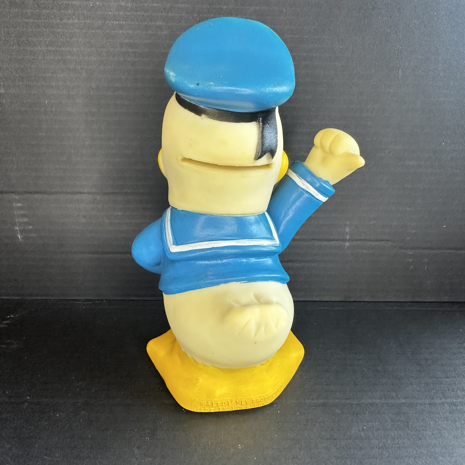 1960s soft vinyl Donald Duck coin bank in blue sailor suit, right hand on hip and left arm raised, with coin slot in back of head, showing age-related yellowing and minor paint wear, embossed Walt Disney Productions Hong Kong copyright on base