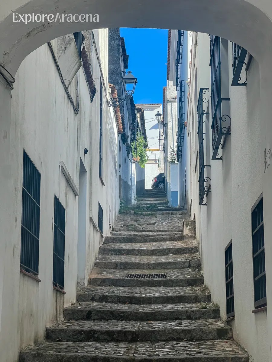 Narrow alleyway in Aracena with stone steps rising uphill between whitewashed buildings and an archway entrance.