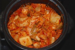Trong một nồi, cho kim chi và thịt heo, bột ớt, tỏi băm, gừng băm vào đun với lửa cho khi kim chi mềm, thịt gần chín (khoảng 10 phút). Thêm nước ép kim chi và nước vào. Giảm nhiệt, đun sôi trong 20 phút, thêm nước nếu cần thiết.
