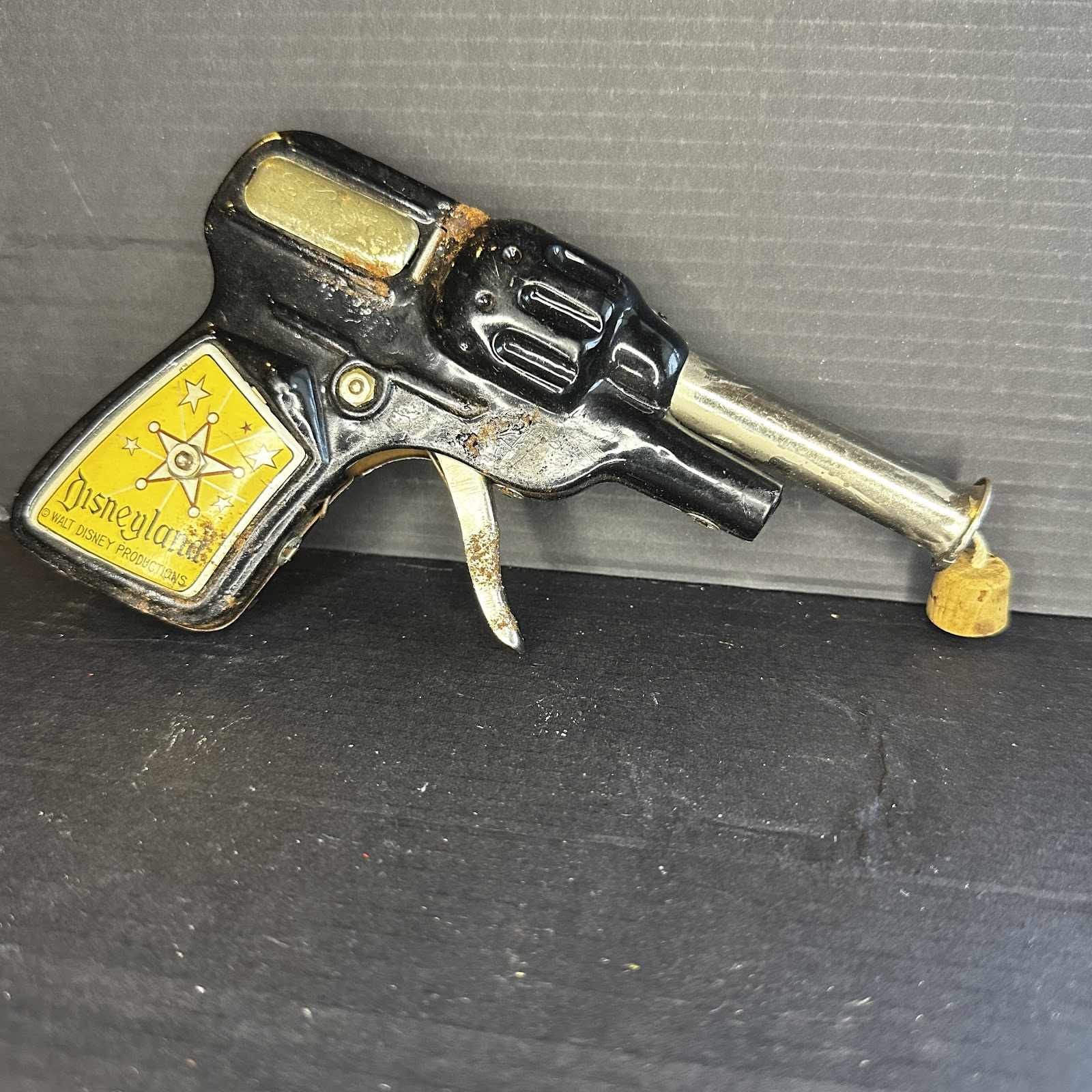 Vintage Disneyland Frontierland cork pop gun in pressed tin with black enamel finish, embossed Disneyland sheriff badge logo, chrome barrel, and original cork on string