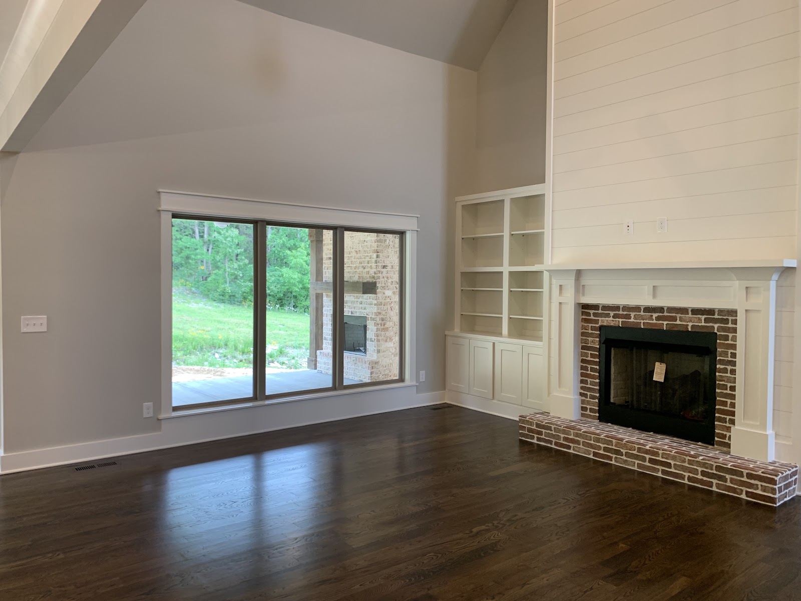 Living room with dark wood floors, a large window overlooking a green landscape, and a brick fireplace with white built-in shelving.