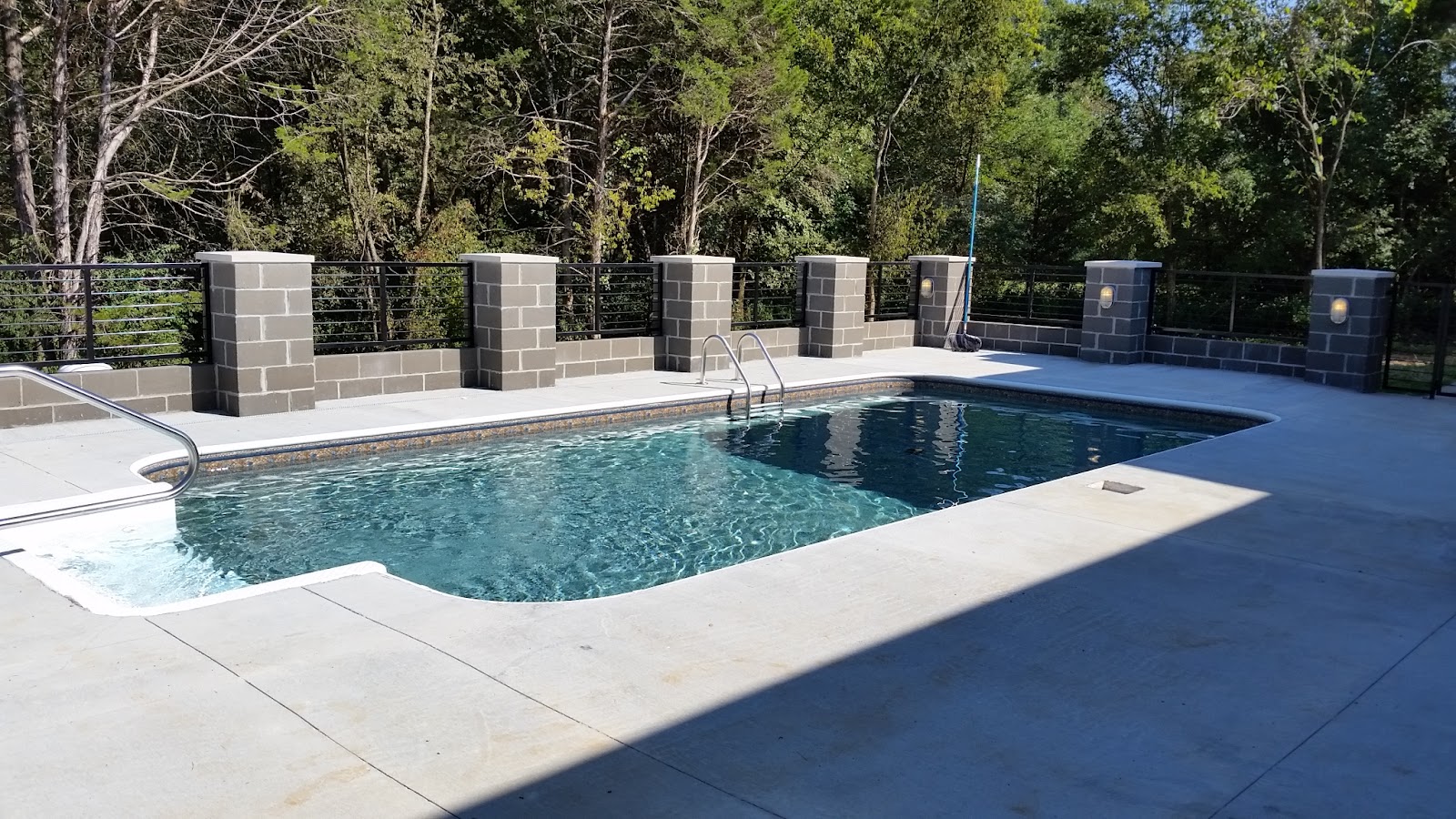 Outdoor swimming pool with a light-colored deck and a block wall fence with dark metal railings.