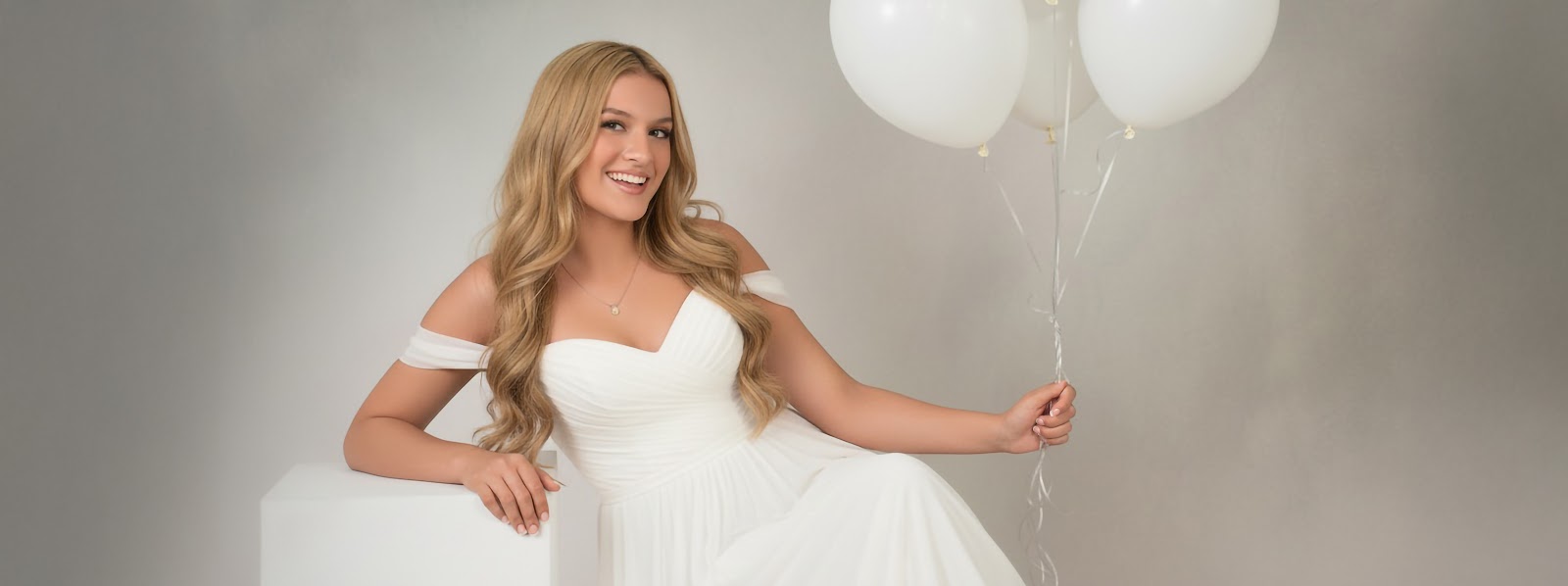 Smiling young woman in an off-shoulder white dress holding white balloons, seated on a cube against a soft studio backdrop.