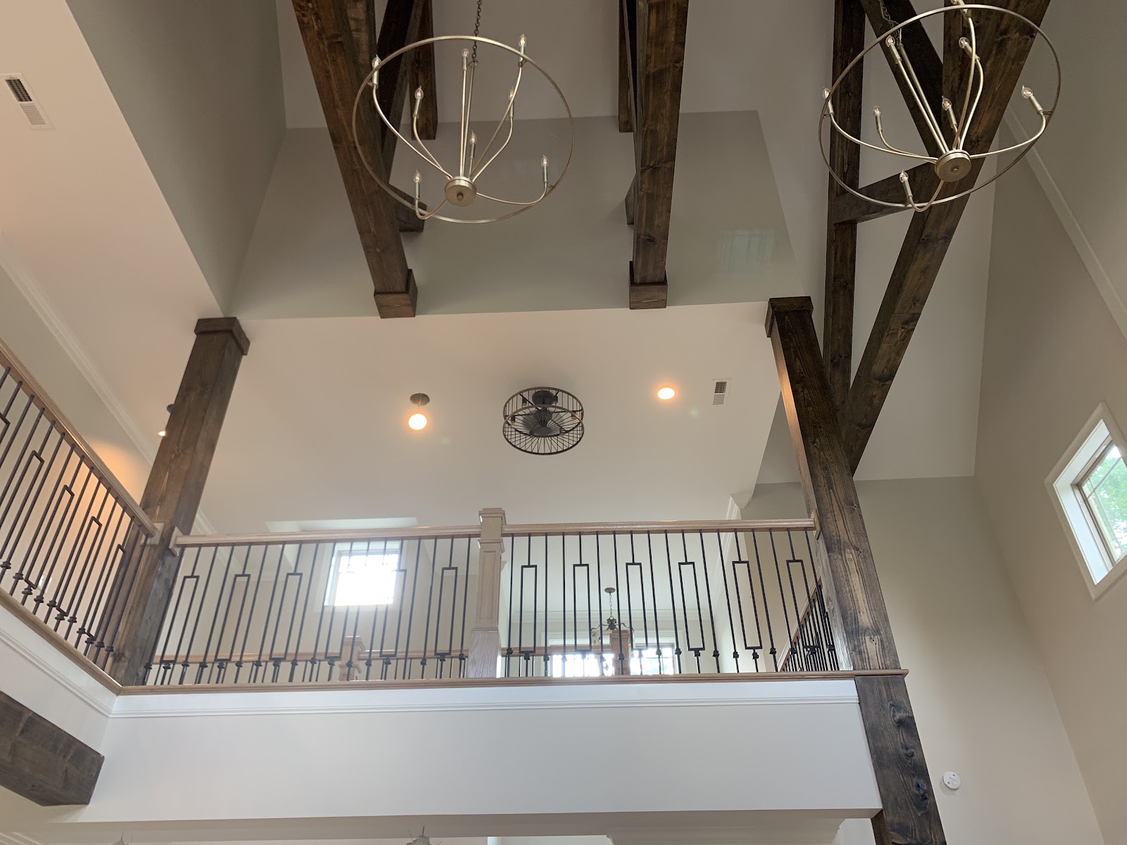 Interior view of a high ceiling with dark wood beams, gold circular chandeliers, and a second-floor balcony with black metal railings.