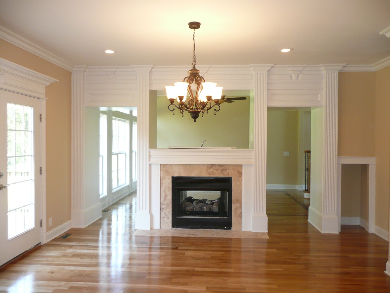 Interior view of a central fireplace with a white mantel and fluted columns, a chandelier, and hardwood floors, with doorways leading to adjacent rooms.