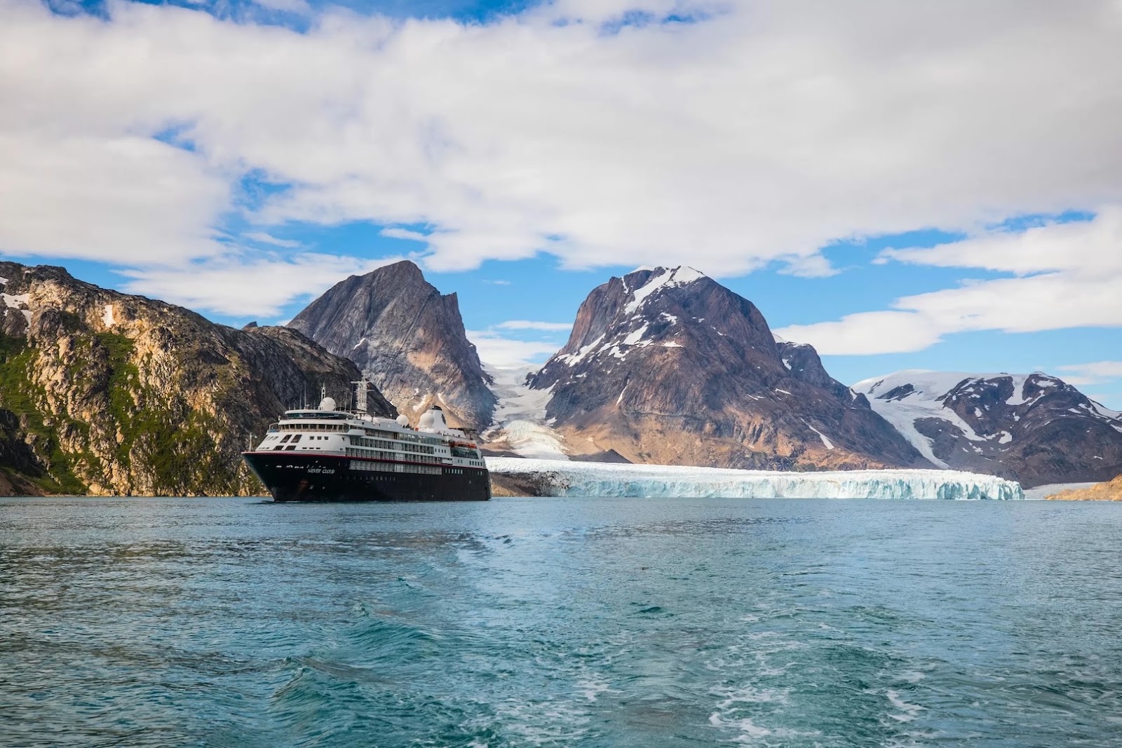 Expedition cruise ship sailing through Greenland waters surrounded by towering icebergs and rugged Arctic landscape.