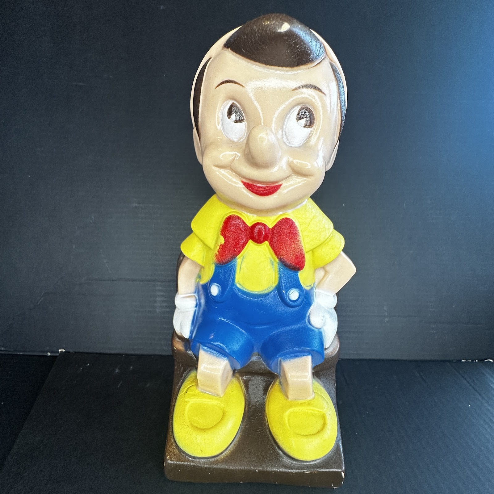 Ceramic chalkware figurine of Pinocchio seated on a brown bench, wearing yellow shirt, blue overalls, red bow tie, and white gloves, with hand-painted details and matte finish