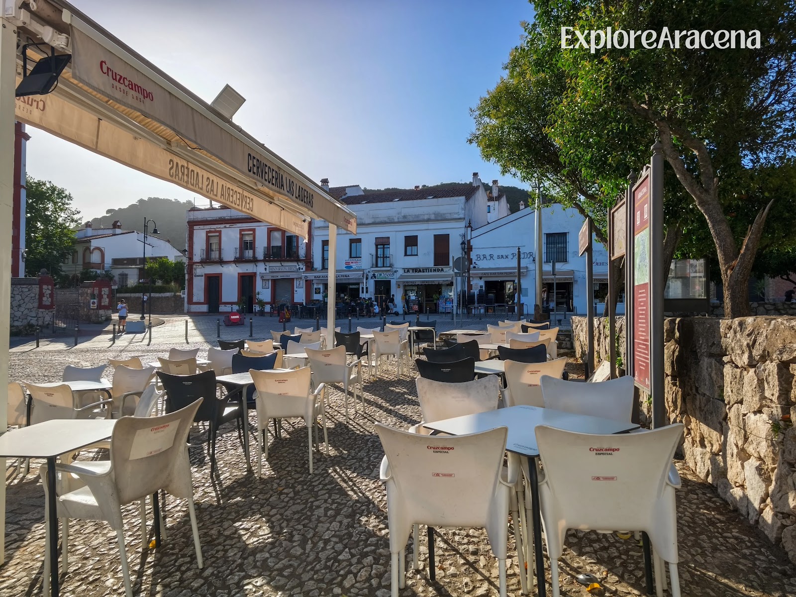 Outdoor tables and chairs beside a bar in a central Aracena plaza during sunset.