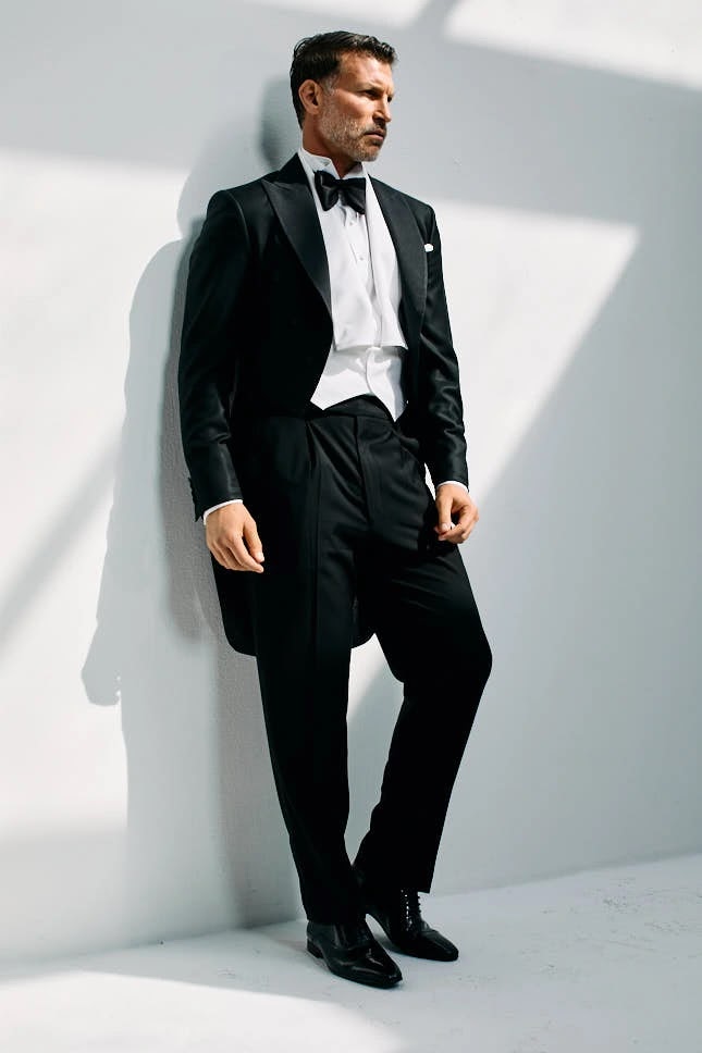 Man in a black tailcoat, white bow tie and waistcoat with patent shoes, full-length against a white wall