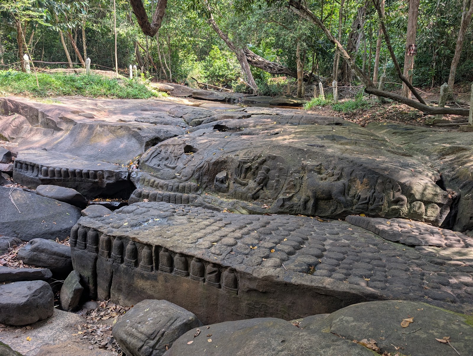 Some large, flat stones. The lowest stone has a series of
                 circles carved into the top face, and some pillar-like
                 structures carved into the side. A neighboring stone has some
                 people and a cow or horse carved into the side. A very slight
                 trickly can be seen moving between some of the stones.