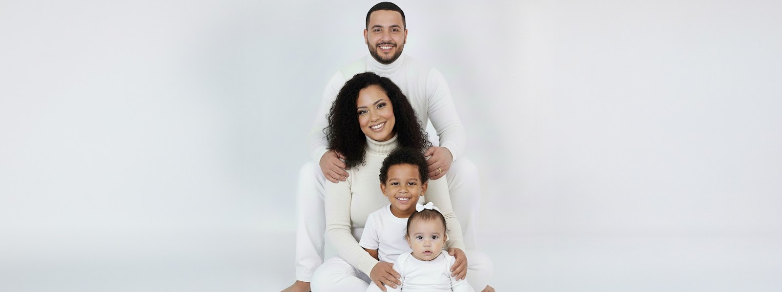 Family of four in a stacked pose on a white studio background—dad behind mom, smiling toddler and baby in front, all in white outfits.