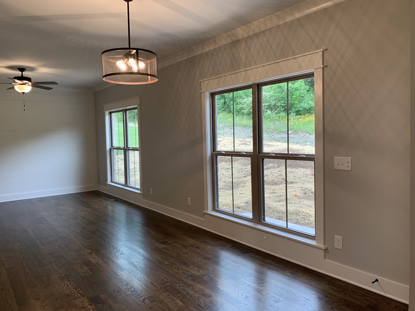 Interior room with dark wood floors and two large windows overlooking a rural landscape. A drum-style chandelier and a ceiling fan are mounted to the ceiling.