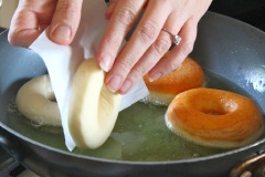 Đặt từng chiếc vòng bánh lên miếng giấy nến hình vuông (mỗi bánh 1 miếng giấy nhé), bạn chờ cho donut nở lớn thêm nữa là có thể chiên nhé. Chế dầu ngập chảo, đun nóng, sau đó cho từng chiếc bột bánh vào chiên đến khi thấy bánh chín, có màu vàng nâu đậm, giòn rụm thì vớt ra cho ráo dầu. Lăn từng chiếc bánh qua một lớp đường để chúng phủ đều bên ngoài là xong rồi đó.
