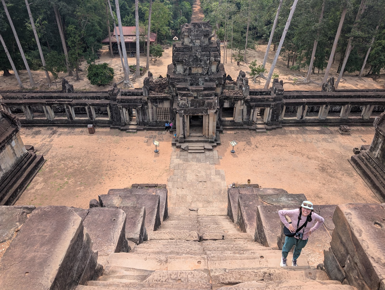 A photo looking down a steep set of stairs showing Kaila on
                 the right side of the staircase. The bottom of the stairs
                 show a plaza and a towering stone gate through an external
                 wall