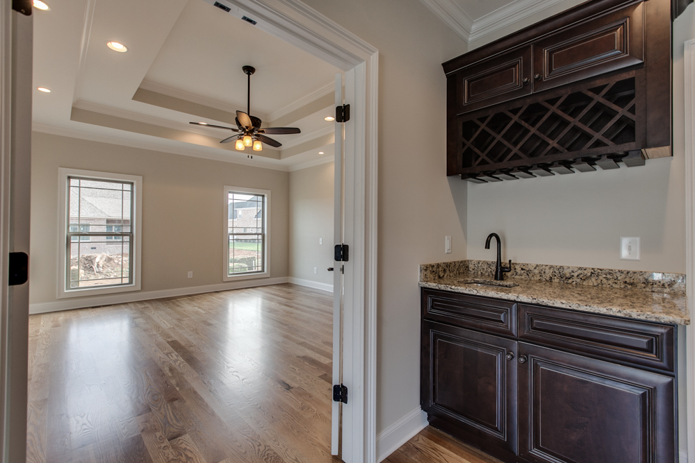 A wet bar with dark wood cabinets and a wine rack, viewed through a doorway leading to a room with hardwood floors and a ceiling fan.