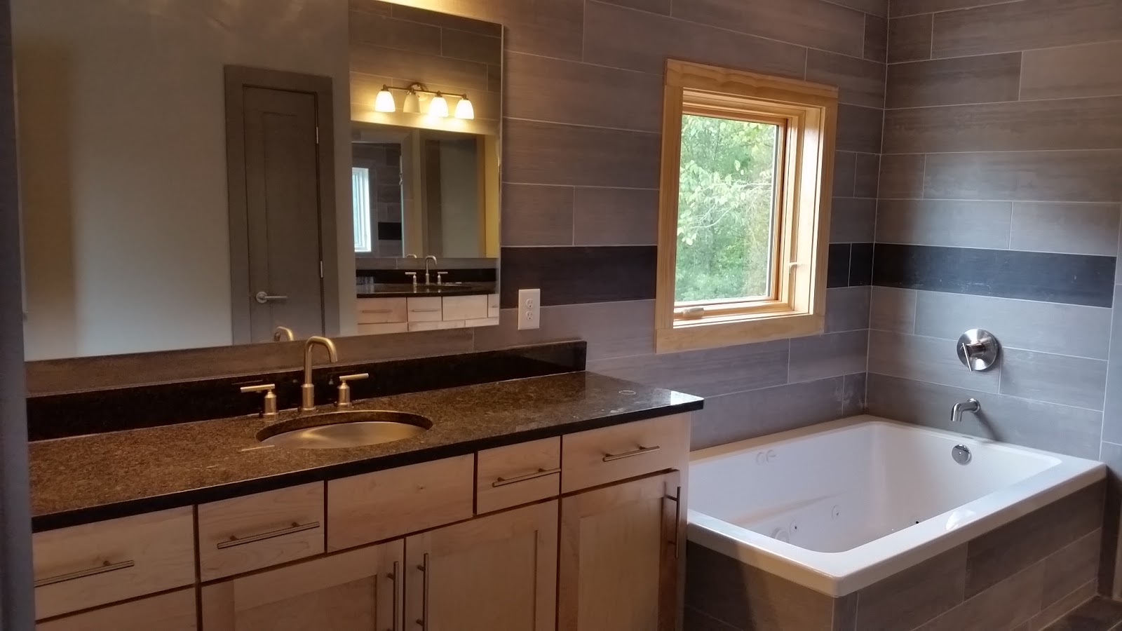 Bathroom interior with a double vanity, dark countertop, light wood cabinetry, and a white bathtub, set against grey tiled walls in East Tennessee.