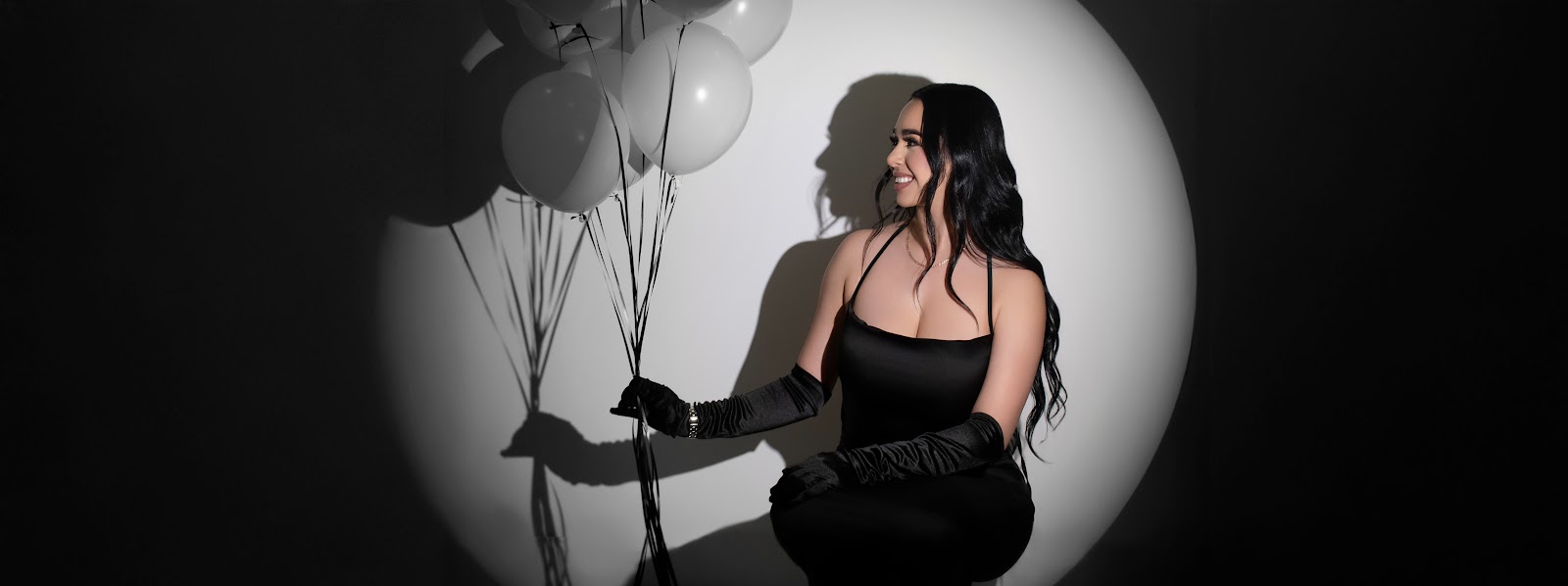 Woman in a black dress and long gloves holding balloons, lit by a circular spotlight with strong shadows on a dark studio backdrop.