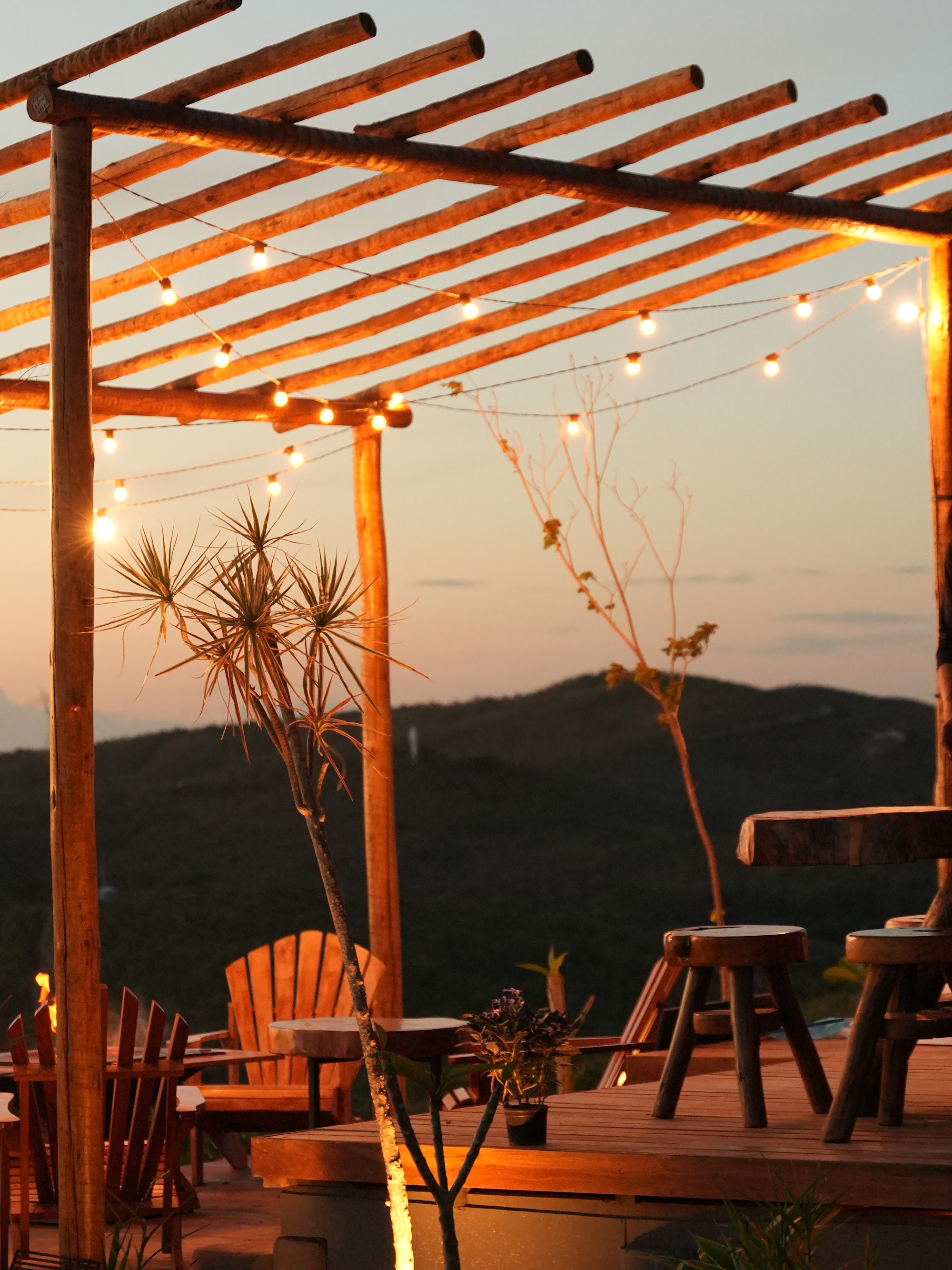 Pergola de madeira com luzes de varal acesas ao entardecer, mesas e cadeiras no deck com vista para as montanhas da Serra do Cipó