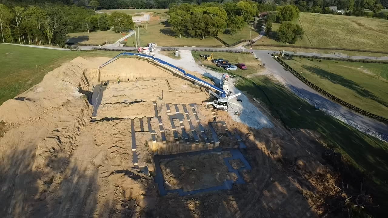 Aerial view of a construction site showing a concrete pump truck pouring foundation footings in an excavated dirt lot.
