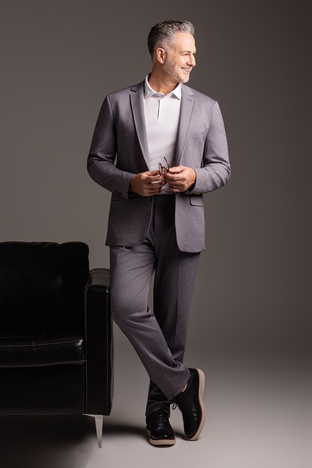 Homem em terno cinza ao lado de poltrona preta em campanha de moda masculina.
