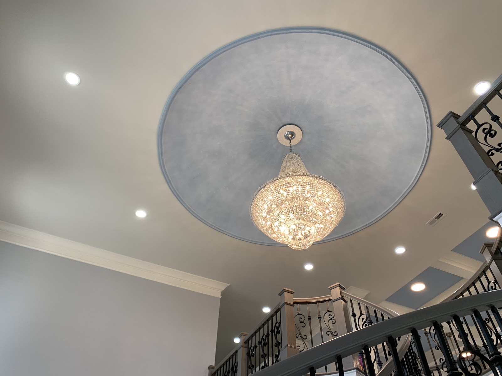A crystal chandelier hangs from a circular blue recessed ceiling feature in a residential interior. Recessed lighting and a staircase with dark metal railings are visible.