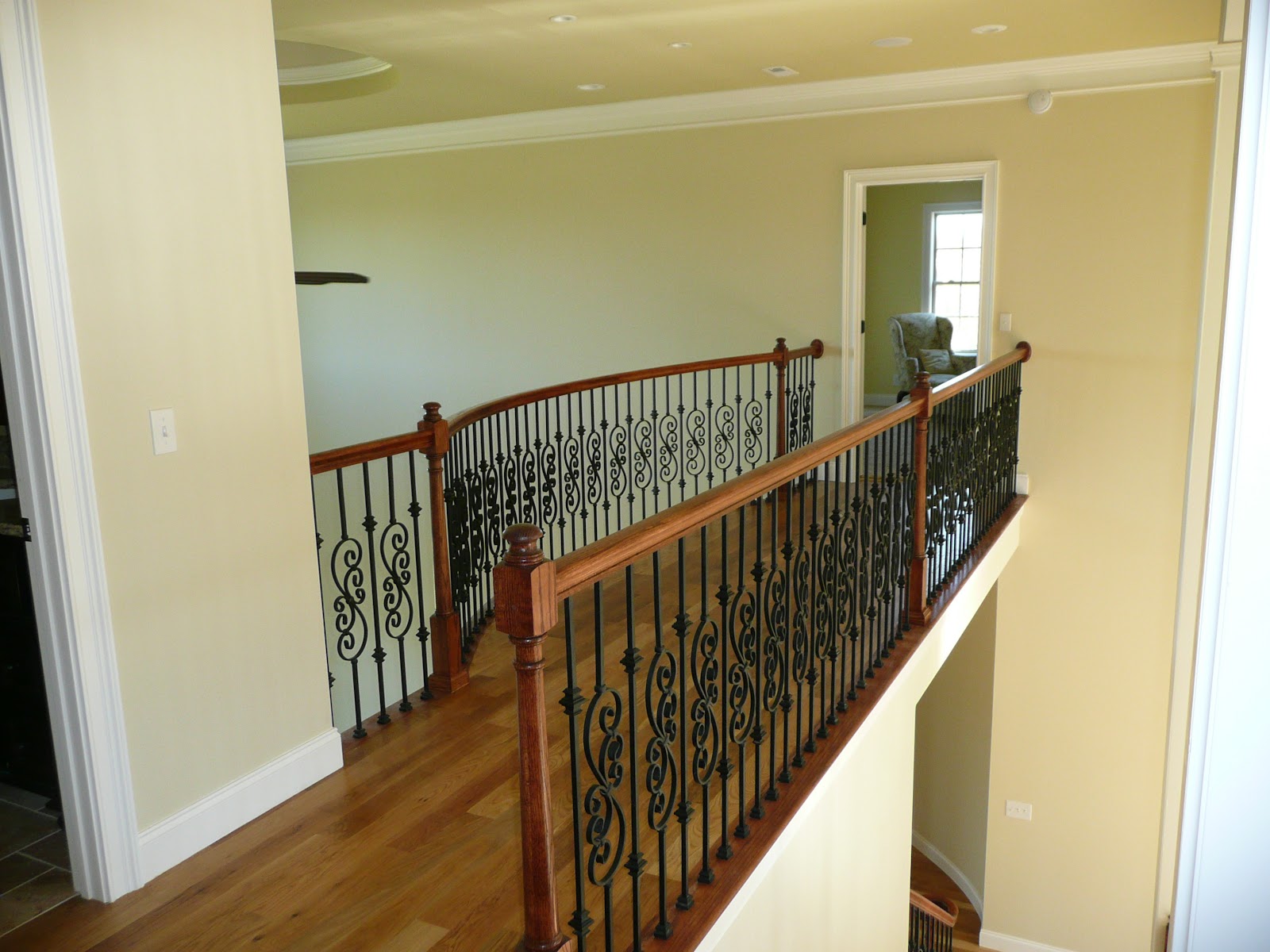 Upper-level hallway with hardwood flooring and a curved wooden railing featuring decorative black metal balusters.