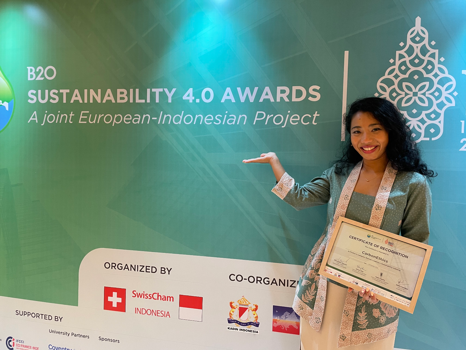 Innandya Kusumawardhani van der Kolk posing in front of B20 Sustainability 4.0 Awards A joint European-Indonesian Project banner Banner holding a certificate of recognition