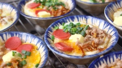 Chuẩn bị bếp nướng than. Đặt từng chén sành lên bếp, cho 1 cục bơ lạt vào chén, tiếp theo đập 4 trứng cút, lần lượt thêm các topping nướng kèm như thịt xay, xúc xích, phô mai, rắc thêm hành lá cắt nhỏ và hành phi. Nướng đến khi từng chén trứng sôi ùng ục, chín mềm thơm bên trong, bên ngoài có lớp vỏ vàng giòn.