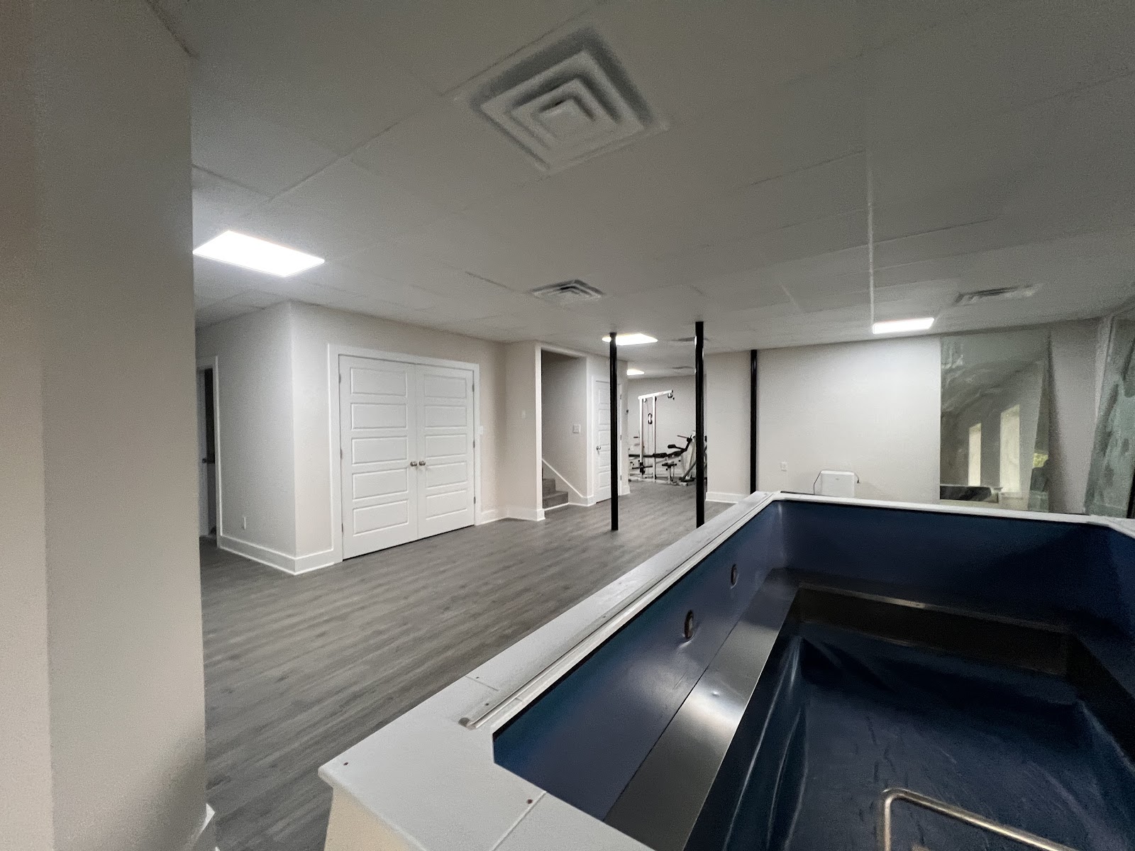 Interior view of a large room featuring dark wood-look flooring and a blue rectangular basin in the foreground. White double doors and an open doorway are visible in the background under a drop ceiling.