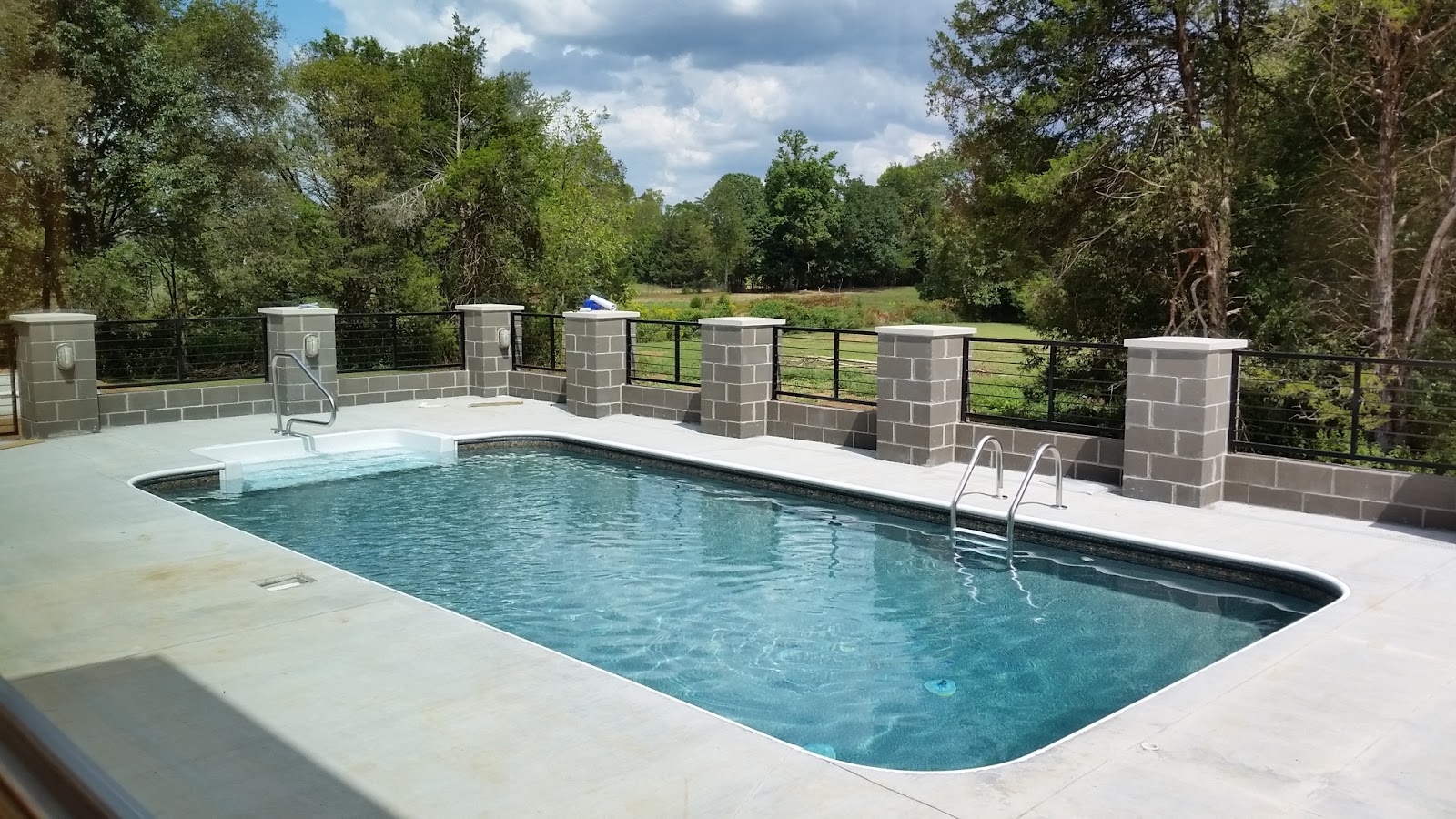 Outdoor swimming pool area with a light-colored deck and a block wall fence under a cloudy sky.