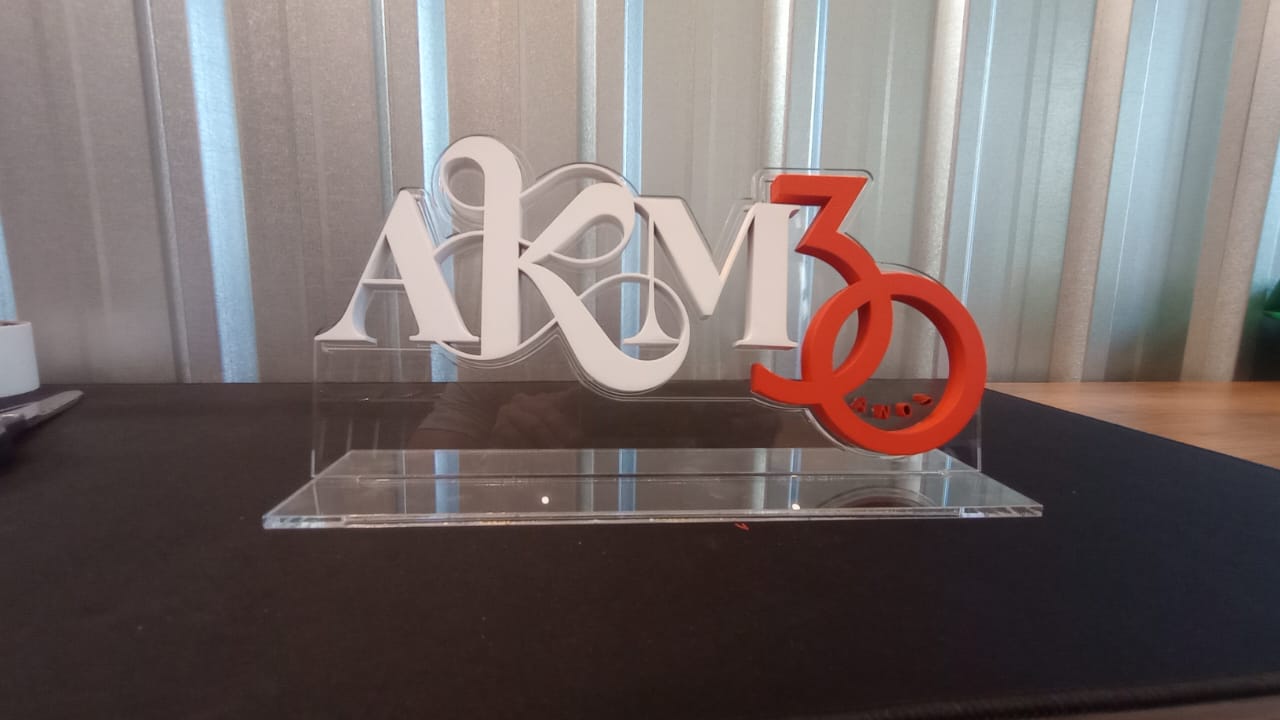 Letreiro AKM 30 finalizado, com letras brancas e números laranja sobre base de acrílico, em uma mesa.